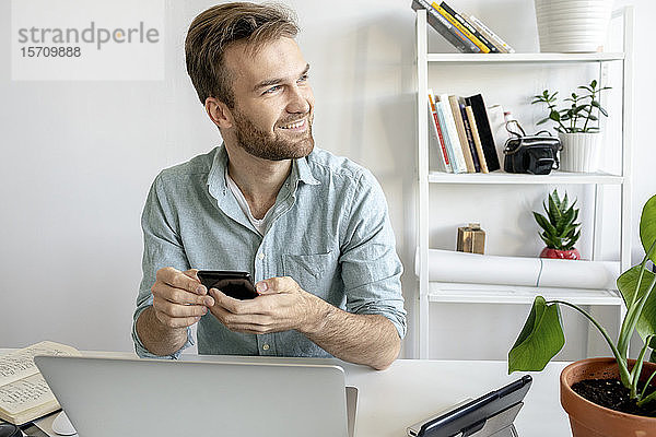 Lächelnder Mann mit Smartphone am Schreibtisch im Büro schaut zur Seite