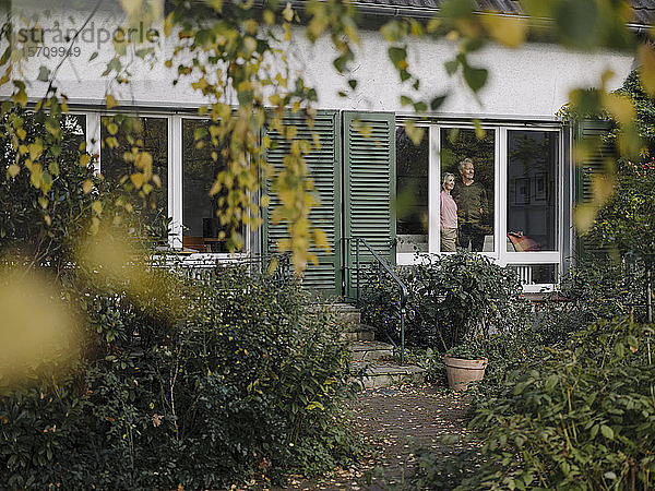 Älteres Ehepaar hinter der Fensterscheibe seines Hauses mit Blick nach draußen