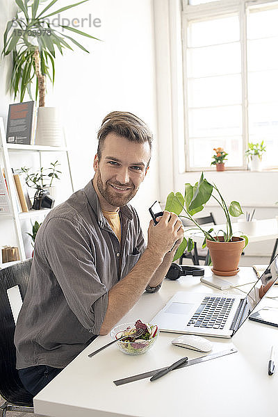 Porträt eines lächelnden Mannes mit Smartphone in der Mittagspause am Schreibtisch im Büro