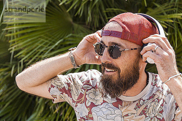 Lächelnder reifer Mann mit roter Basecap  Sonnenbrille und weißen Kopfhörern