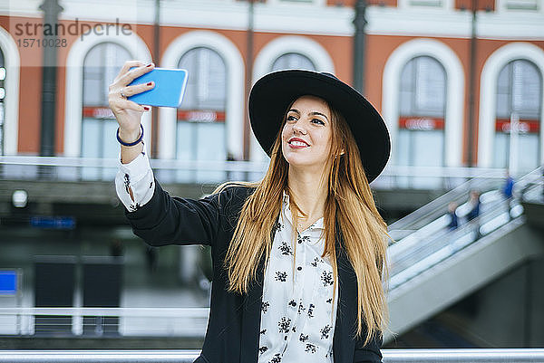 Lächelnde junge Frau mit Hut nimmt am Bahnhof ein Selfie