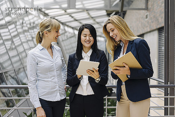 Drei Geschäftsfrauen mit Tablett sprechen in modernem Bürogebäude