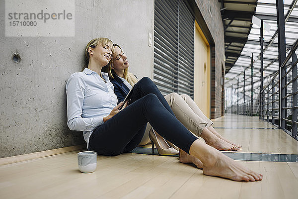 Zwei junge Geschäftsfrauen machen eine Pause im Büro und sitzen auf dem Boden