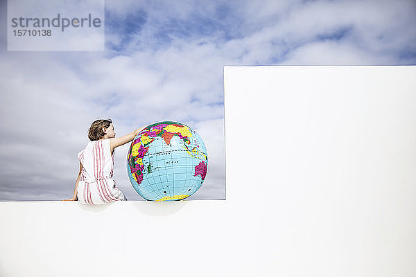Kleines Mädchen an der Wand sitzend  mit Arm um den aufblasbaren Globus  Rückansicht