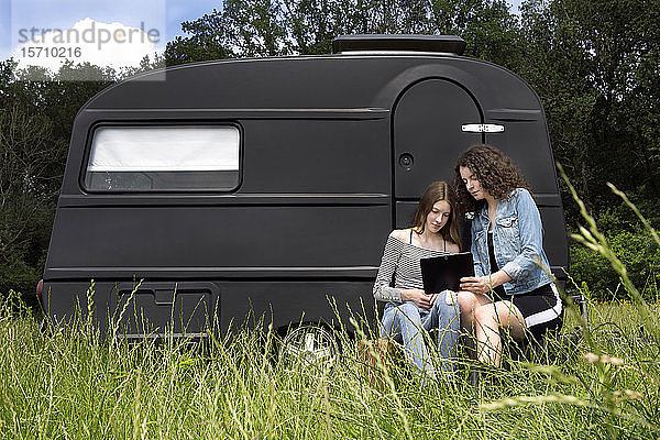 Zwei Freunde sitzen auf einer Wiese vor einem schwarzen Wohnwagen und benutzen ein digitales Tablett