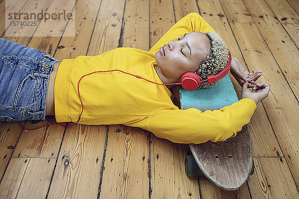 Junge Frau liegt zu Hause auf dem Skateboard auf dem Boden und hört Musik