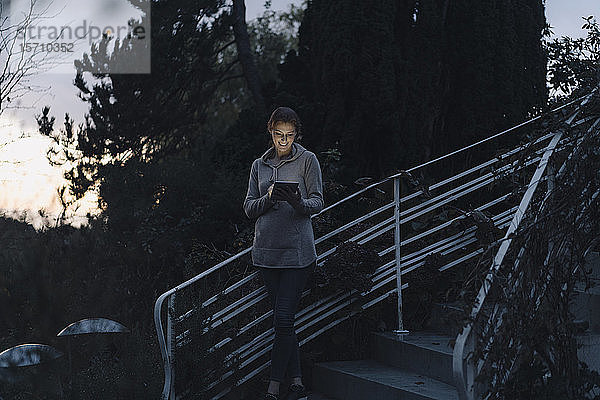 Frau benutzt digitales Tablett in der Dämmerung  steht auf einer Treppe im Garten