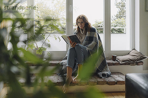 Eine reife Frau sitzt auf einer wunderbaren Fensterbank  in eine Decke gewickelt  liest ein Buch
