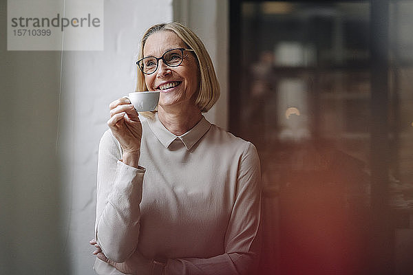 Lächelnde reife Geschäftsfrau macht im Büro eine Kaffeepause am Fenster
