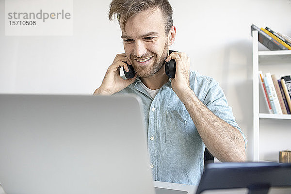 Lächelnder Mann mit Kopfhörern und Laptop am Schreibtisch im Büro