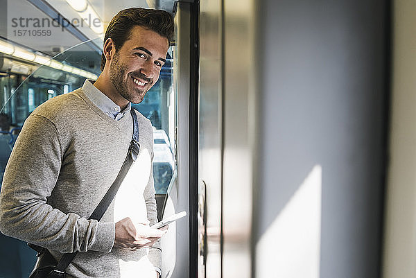 Porträt eines lächelnden jungen Mannes mit Smartphone in einem Zug