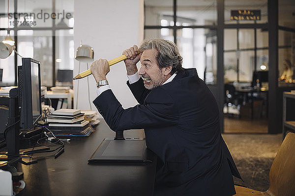 Verärgerter leitender Geschäftsmann mit riesigem Bleistift am Schreibtisch im Büro