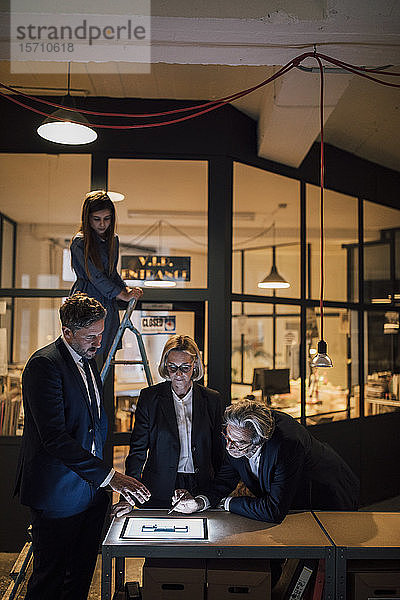 Geschäftsleute diskutieren an glänzender Tafel im Büro mit daneben stehendem Mädchen auf Leiter