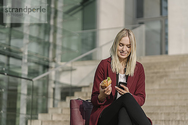 Blonde Geschäftsfrau isst draußen zu Mittag und benutzt ein Smartphone