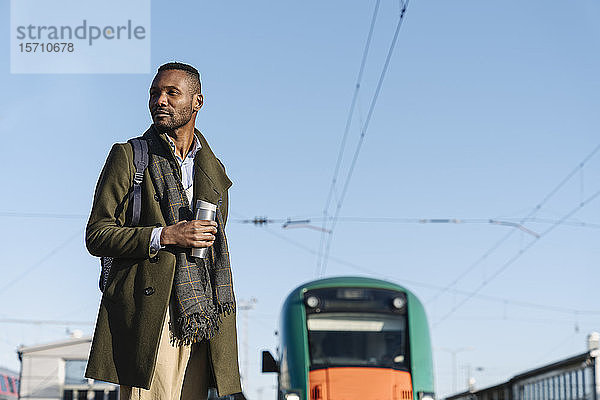 Porträt eines stilvollen Mannes mit wiederverwendbarem Becher  der auf den Zug wartet