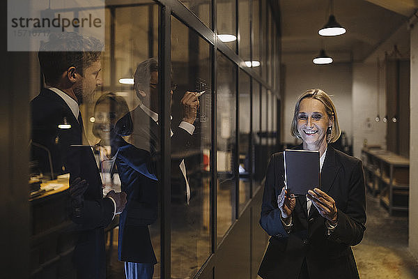 Lächelnde Geschäftsfrau mit Tablett und zwei Geschäftsleute  die im Büro an einer Glasscheibe zeichnen