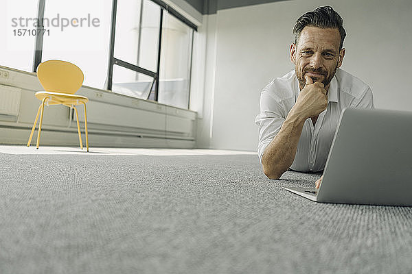 Porträt eines reifen Geschäftsmannes  der in einem leeren Büro auf dem Boden liegt und einen Laptop benutzt