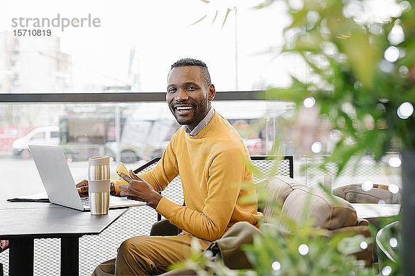 Porträt eines glücklichen Mannes mit Smartphone und Laptop in einem Cafe