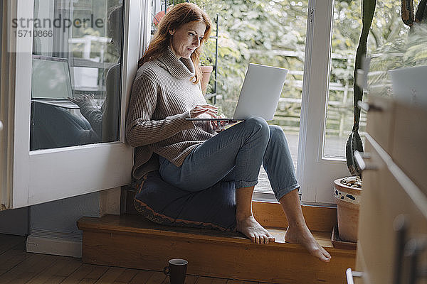 Eine reife Frau sitzt auf den Stufen einer Balkontür und benutzt einen Laptop