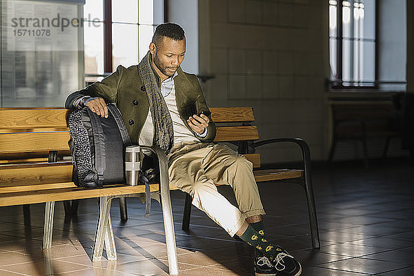 Stylischer Mann benutzt Smartphone  während er auf einer Bank in einem Bahnhof sitzt