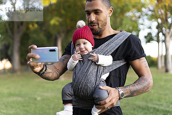 Junger Vater trägt seinen kleinen Sohn im Tragetuch und nimmt Smartphone-Selfie