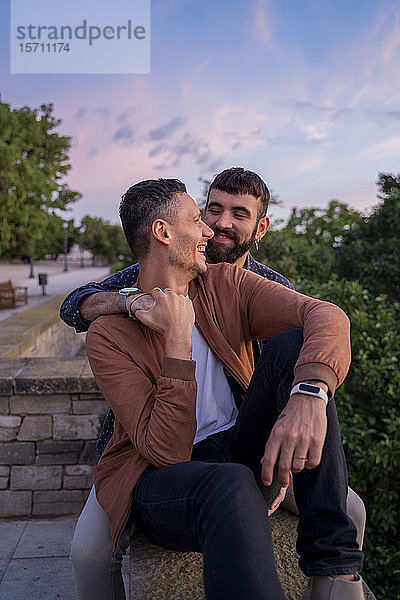 Glückliches  anhängliches schwules Paar sitzt bei Sonnenuntergang auf einer Mauer