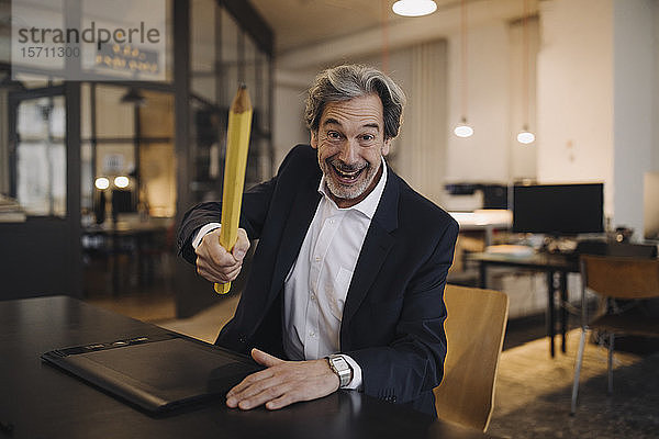 Dummer leitender Geschäftsmann mit riesigem Bleistift am Schreibtisch im Büro
