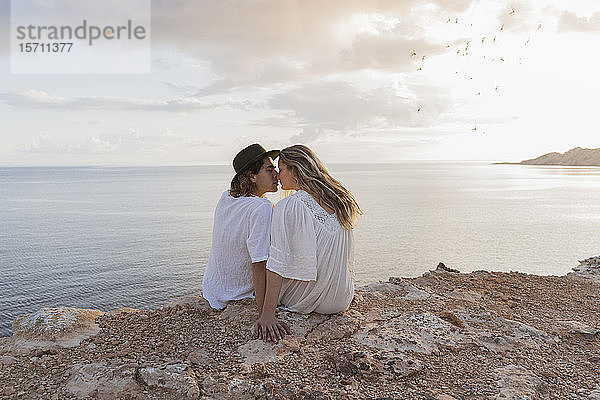 Rückenansicht eines jungen verliebten Paares  das auf einem Felsen vor dem Meer sitzt  Ibiza  Balearen  Spanien
