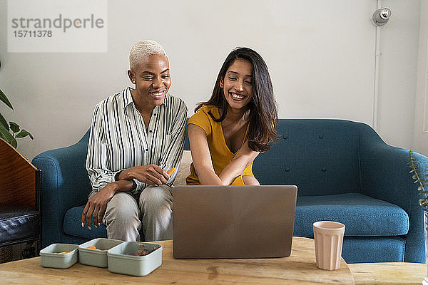 Zwei glückliche Frauen sitzen zusammen auf der Couch und benutzen einen Laptop