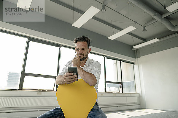 Reifer Geschäftsmann sitzt auf gelbem Stuhl in leerem Büro und benutzt Smartphone