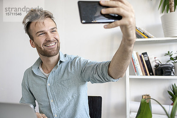 Lächelnder Mann nimmt sich am Schreibtisch im Büro ein Selfie