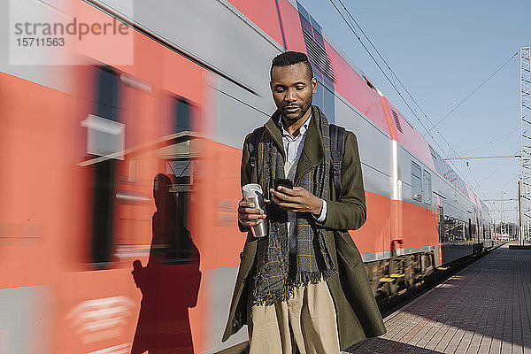 Porträt eines stilvollen Mannes  der sein Smartphone neben einem Zug benutzt