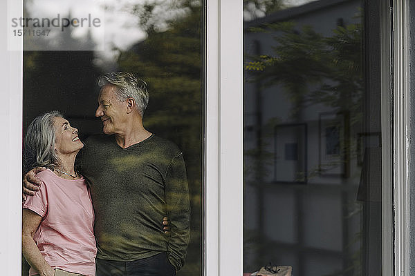 Glückliches älteres Ehepaar hinter der Fensterscheibe ihres Hauses