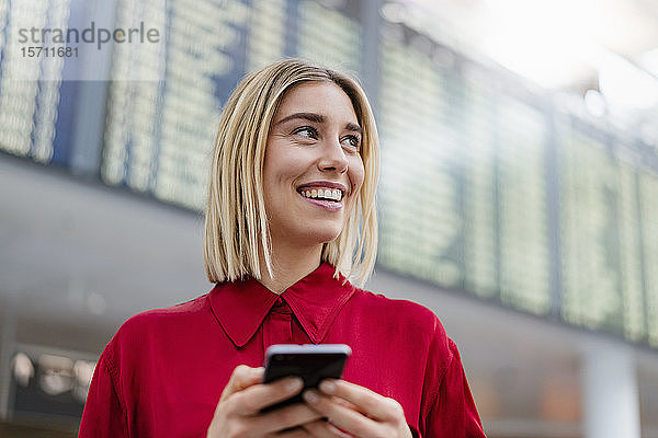 Lächelnde junge Geschäftsfrau mit Handy bei der Ankunft Abflugtafel am Flughafen