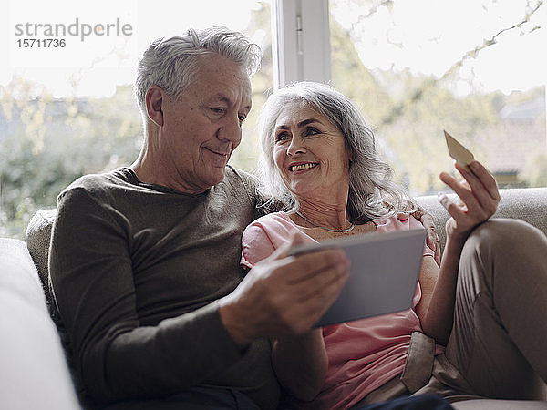 Glückliches älteres Ehepaar entspannt sich zu Hause auf der Couch und nutzt Tablet zum Online-Shopping