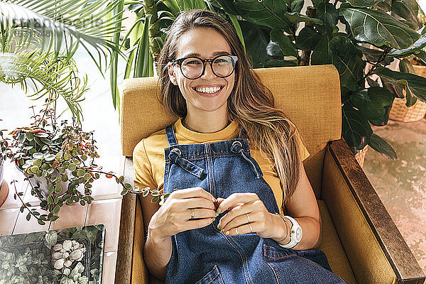 Porträt einer glücklichen jungen Frau im Sessel sitzend  umgeben von Pflanzen