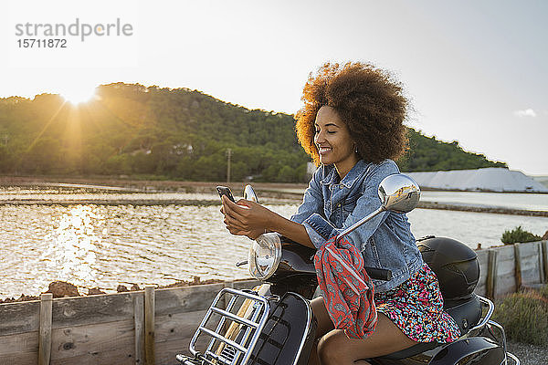 Junge Frau sitzt auf einem Roller und benutzt ein Smartphone bei Sonnenuntergang  Ibiza