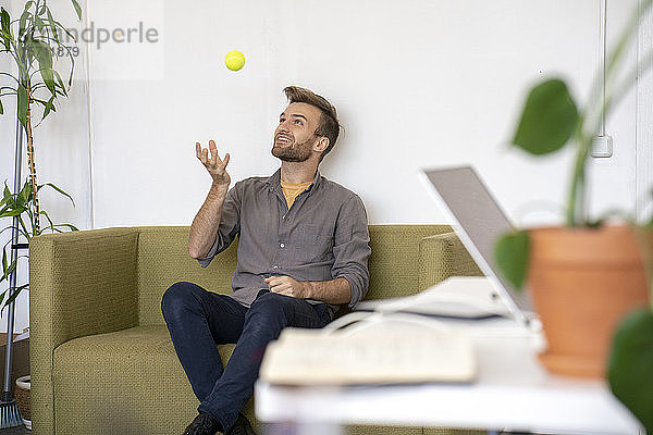 Lächelnder Mann sitzt im Büro auf der Couch und spielt mit einem Tennisball