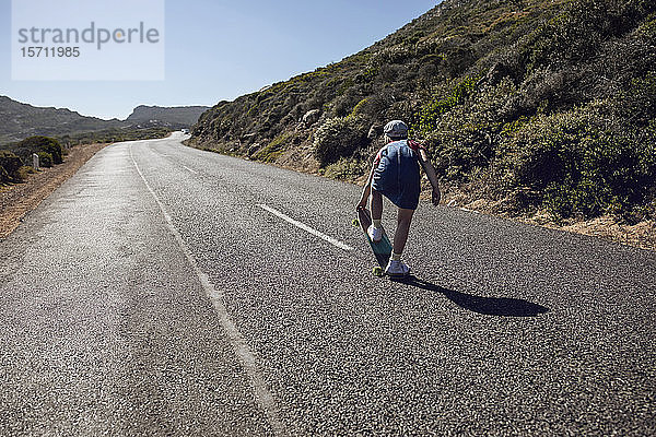 Rückenansicht eines Mädchens mit Skateboard auf der Landstraße stehend  Kapstadt  Western Cape  Südafrika