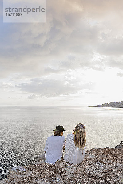 Rückenansicht eines jungen Paares  das auf einem Felsen vor dem Meer sitzt  Ibiza  Balearen  Spanien