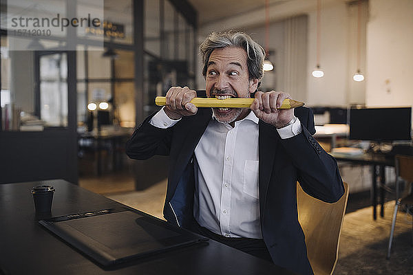 Verärgerter leitender Geschäftsmann mit riesigem Bleistift am Schreibtisch im Büro