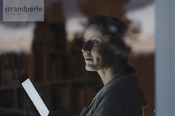 Frau  die in der Dämmerung aus dem Fenster schaut und ein digitales Tablet hält