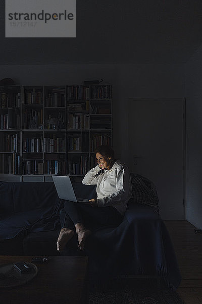 Frau sitzt auf der Couch eines dunklen Wohnzimmers und benutzt einen Laptop