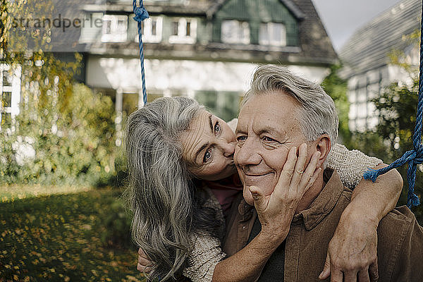 Glückliche Frau umarmt einen küssenden älteren Mann auf einer Schaukel im Garten