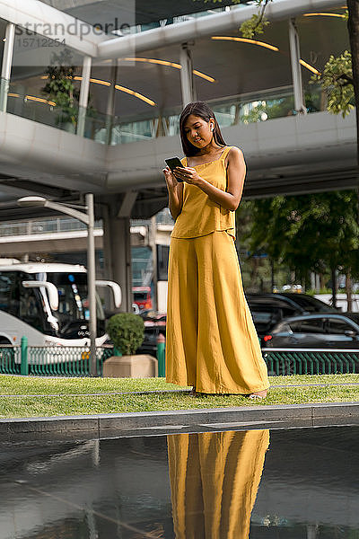 Porträt einer modischen  gelb gekleideten Frau mit Ohrstöpseln und Smartphone in der Stadt