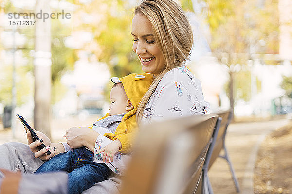 Glückliche Mutter ruht sich mit ihrem kleinen Jungen auf einer Parkbank aus und benutzt ein Smartphone