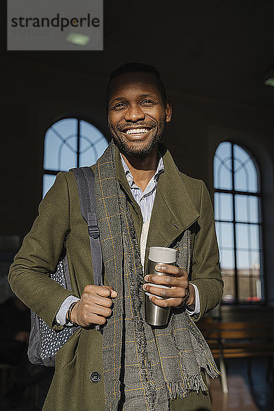 Porträt eines glücklichen stilvollen Mannes mit wiederverwendbarer Tasse im Bahnhof
