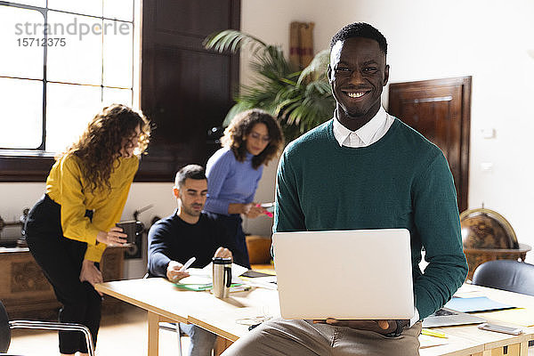Porträt eines lächelnden Mannes im Büro mit Laptop und Kollegen im Hintergrund