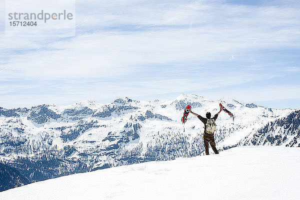 Mann mit Rucksack  auf Berggipfel stehend  winkt mit seinen Schneeschuhen  Rückansicht  Bundesland Salzburg  Österreich