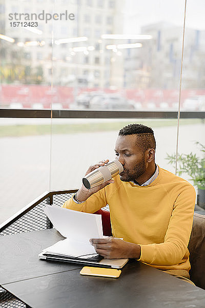 Mann trinkt heißes Getränk aus einem wiederverwendbaren Becher  während er in einem Café Dokumente liest